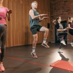 Four adults performing high knees in an indoor gym with wooden panel walls.
