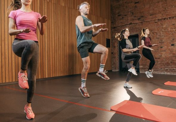 Four adults performing high knees in an indoor gym with wooden panel walls.