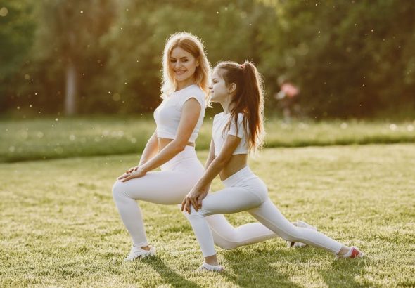 A woman and young girl in white sportswear doing stretches on a grassy field under the sunlight.