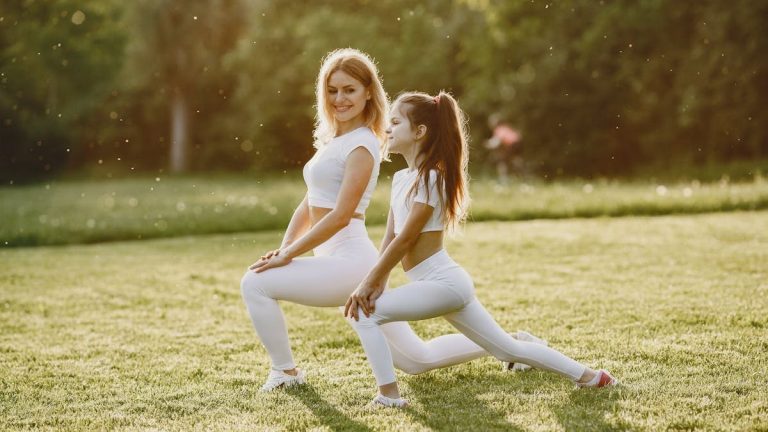 A woman and young girl in white sportswear doing stretches on a grassy field under the sunlight.